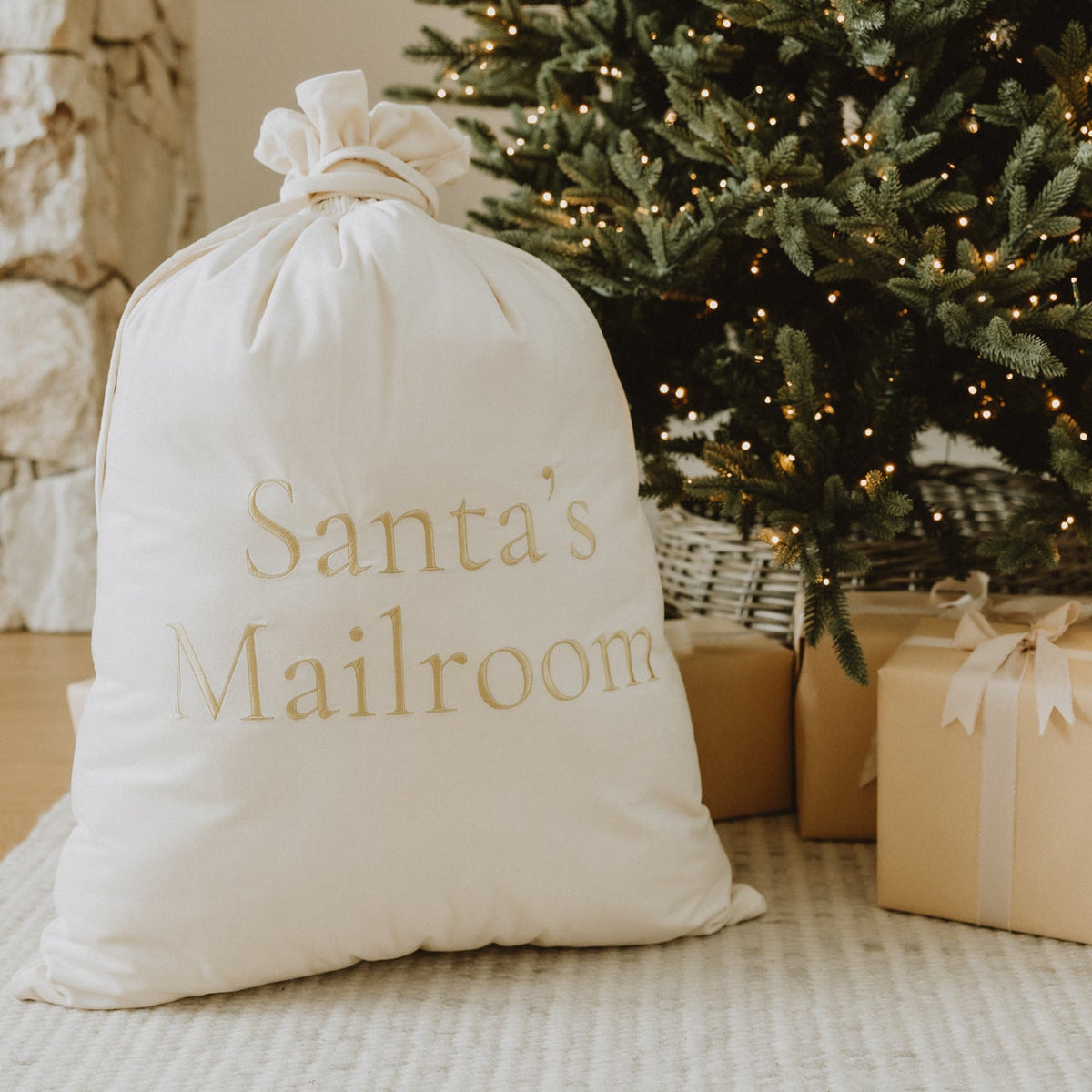 A white fabric Santa mailer bag with gold lettering reading 'Santa's Mailroom' sits on a light rug beside wrapped gift boxes and a decorated Christmas tree in a cozy indoor setting.