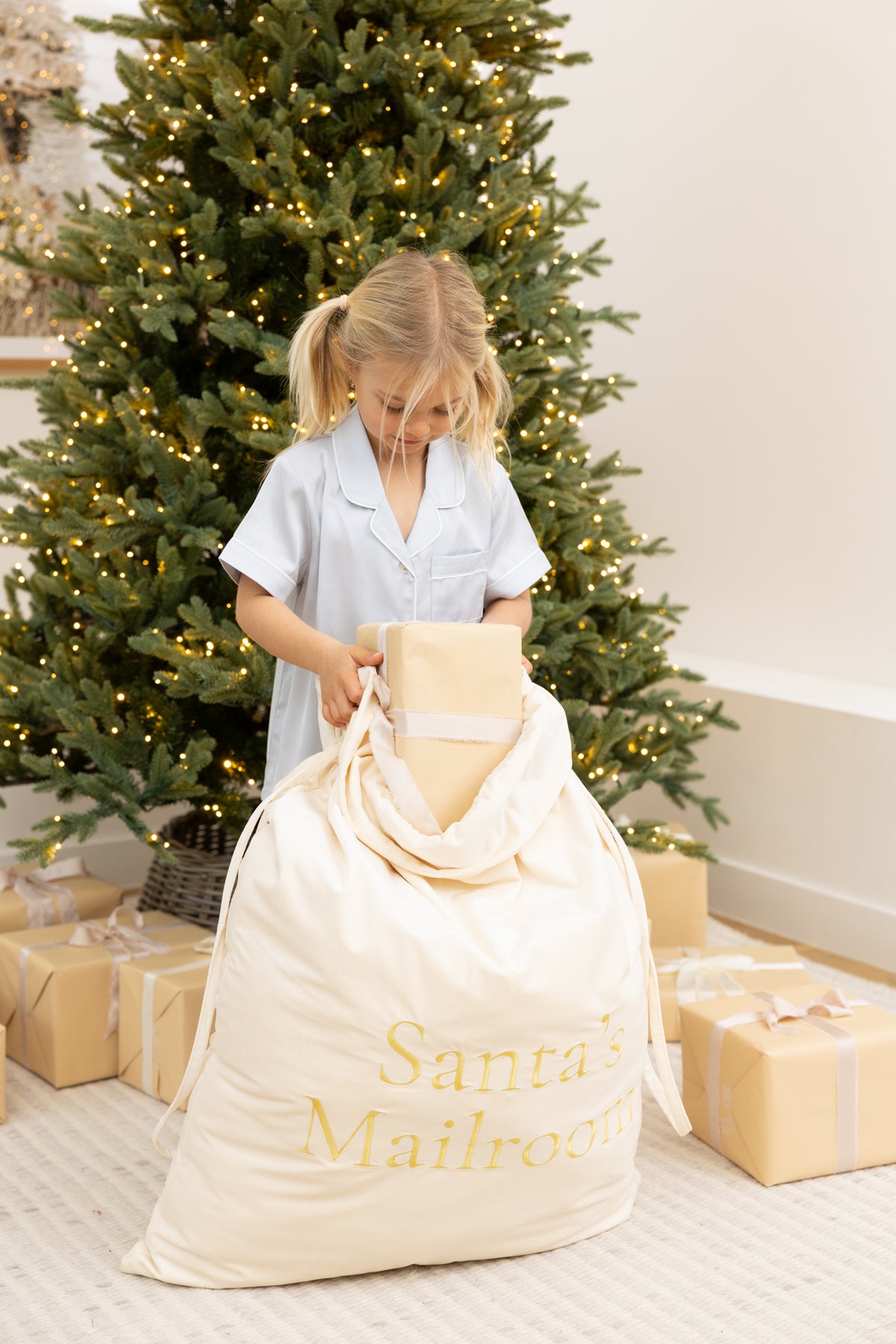 A young girl in a light blue pajama shirt stands in front of a decorated Christmas tree, holding a large cream-colored Santa's Mailroom sack with gold lettering. 