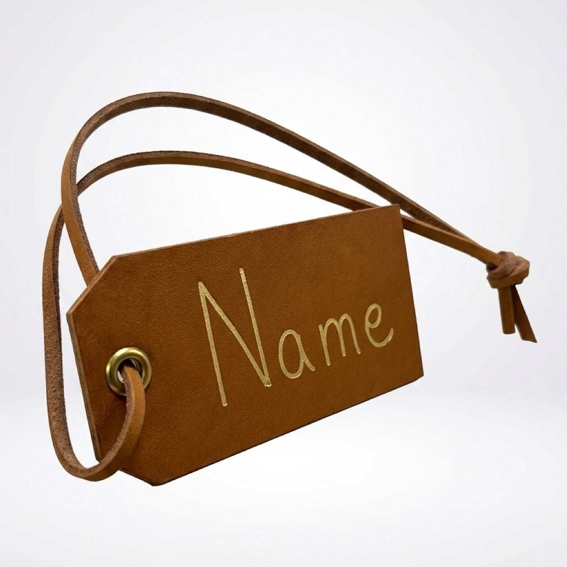 A brown faux leather luggage tag with a matching leather loop strap and a gold-colored metal grommet, featuring the word 'Name' printed in gold on the front, set against a light gray gradient background.