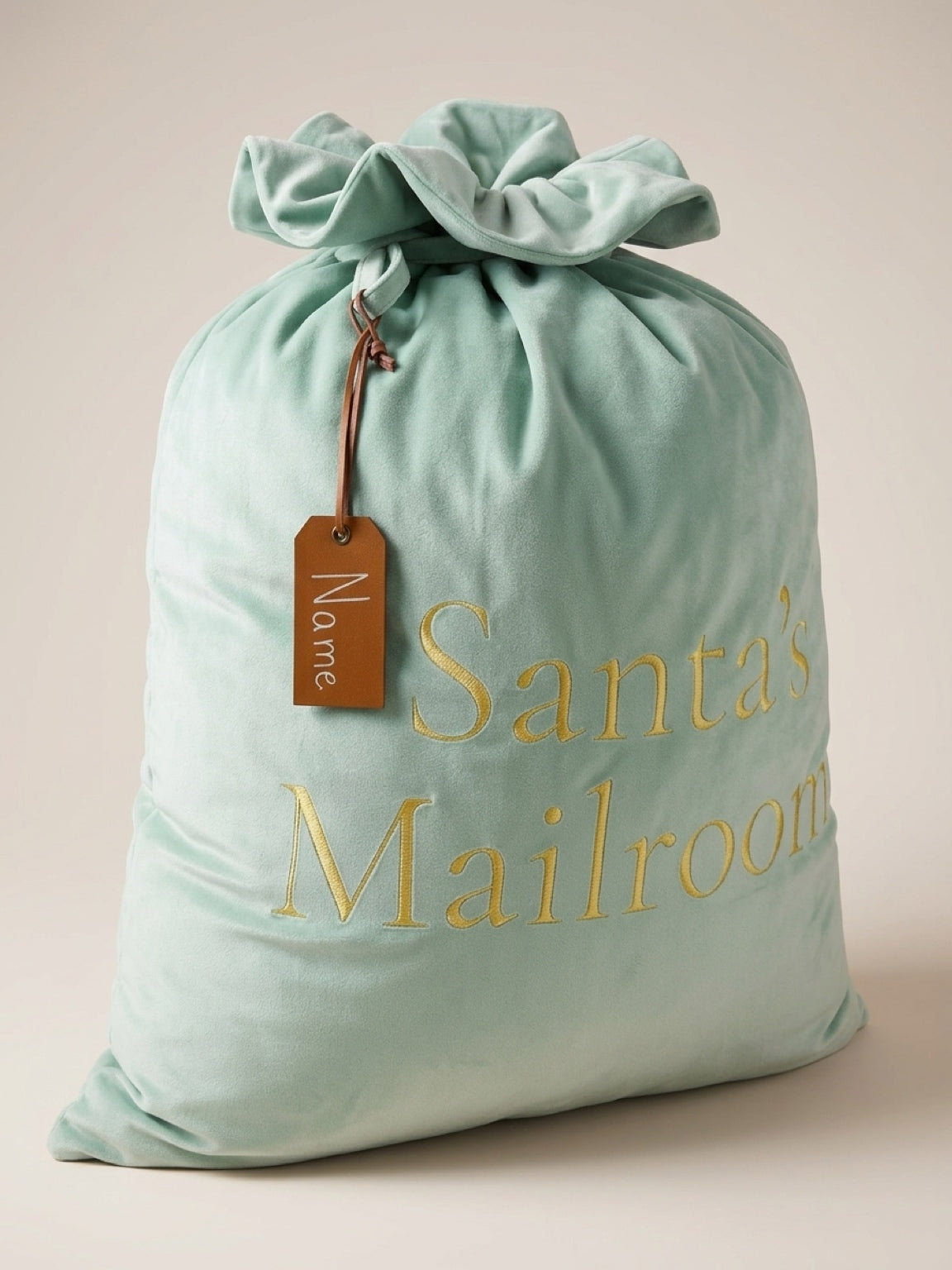 A sage green velvet drawstring Santa mailer bag with gold embroidered text reading 'Santa's Mailroom' and a brown hanging tag labeled 'Name', shown on a plain beige studio background.